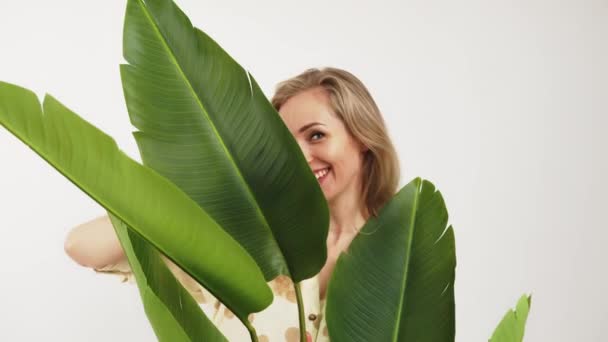 Hermosa risa rubia caucásica joven mujer jugando peekaboo con la cámara, mirando a la cámara desde detrás de las hojas verdes gigantes. Estudio sobre fondo blanco. — Vídeos de Stock