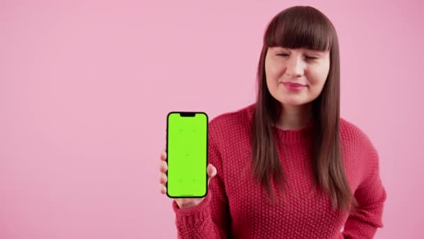 Amicale femme caucasienne tient le téléphone mock up avec écran vert à la caméra, semble surpris, et sourit à la caméra. Espace de copie isolé sur fond rose. — Video