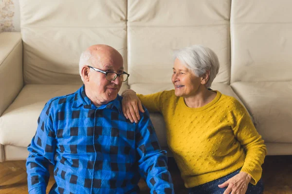 Two pensioner seniors - woman and man - sitting on the floor and leaning their backs on beige leather sofa. Happy retirement concept. — Stockfoto