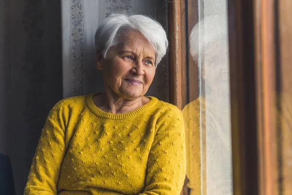 Indoor medium shot of pensive elderly pensioner senior lady looking through the window with soft smile on her lips. — ストック写真