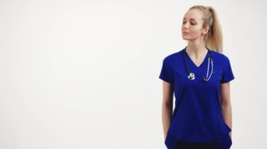 Mavi üniformalı orta boy hemşire ellerini ceplerinde tutuyor ve başını yana çeviriyor.