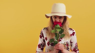 Yakından, elinde güzel kırmızı gül yaprakları tutarken ve koklarken kameraya bakan, şapkalı, çiçekli bir gömlek giyen, sevimli, Avrupalı genç, sarışın kadının portresi..