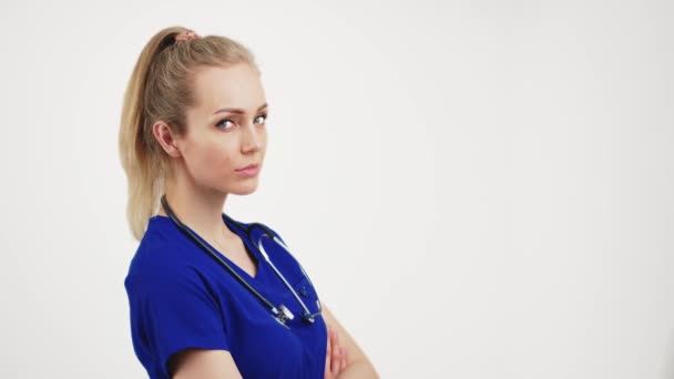 Serious, confident, young caucasian female doctor in blue uniform with stethoscope over her neck in studio over white background. — Video