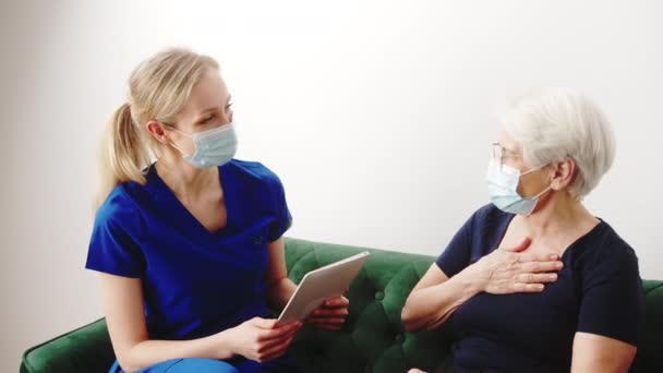 Professionell kvinnlig vårdgivare undersöker senior gråhårig mormor medan du sitter på en mörkgrön soffa. — Stockvideo
