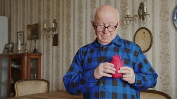 Старший мужчина кладет реалистичное игрушечное сердце в грудь и смотрит в камеру с улыбкой — стоковое видео