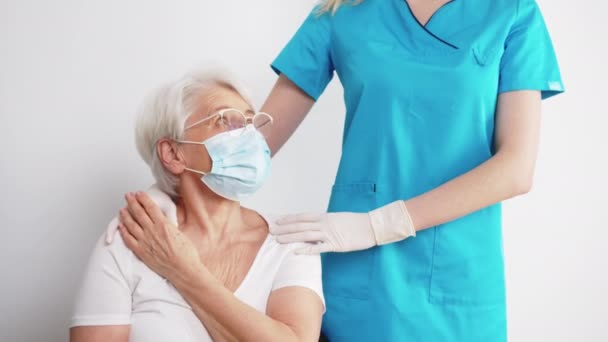 Jovem enfermeira caucasiana em uma roupa azul e luvas de proteção apoia sua senhora paciente idoso em uma máscara, tocando seus braços. Apoio do profissional de saúde. — Vídeo de Stock