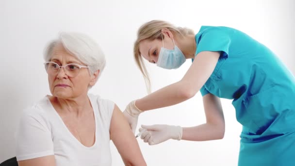 Испуганная пожилая седовласая бабушка в белой футболке получает прививку от медицинского работника в синем костюме. — стоковое видео