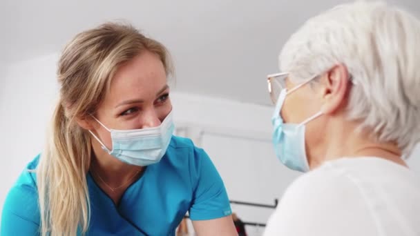 Femme soignante caucasienne masquée avec une pensionnée âgée parlant gaiement. Soutien des travailleurs de la santé. — Video