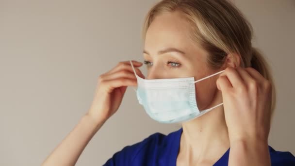 Closeup shot of a caucasian blonde female nurse in a blue uniform putting on a protective face mask. The proper way to put a face mask on. — стоковое видео