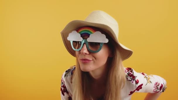Vicces szőke európai nő csitítja a kamerát, miközben vicces szivárvány szemüveget és szalmakalapot visel narancssárga háttér felett. — Stock videók