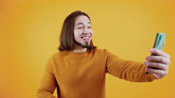 Allegro uomo caucasico barbuto con i capelli lunghi guardando il suo smartphone e parlando con qualcuno via internet chiamata su sfondo giallo. — Video Stock