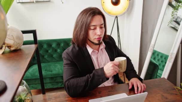 Кавказький позаштатний бізнесмен, який працює з домашнього офісу, сидить за столом, дивлячись на ноутбук, п'є з кавового горнятка, дивлячись суворо на камеру . — стокове відео