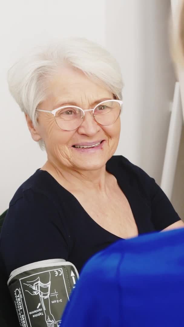 Yaşlı beyaz saçlı kadın gülümseyerek kan basıncını ölçüyor ve dikey video medikal konseptiyle doktorluk yapıyor. — Stok video
