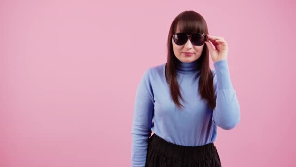 Porträtt av en flicka i blå polotröja på en rosa bakgrund som tar av sig sina solglasögon och ler mot kameran — Stockvideo