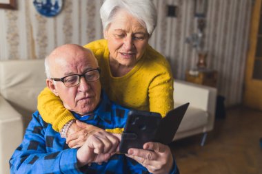 Beyaz saçlı yaşlı kadın emekli kocasına arkadan sarılıyor ve modern akıllı telefonunda ona yardım ediyor. Klasik oturma odası iç mimarisi..
