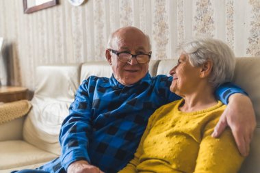 Yaşlı beyaz heteroseksüel çift bej bir deri koltukta oturup birbirlerine sarılıyorlar ve geçmişteki deneyimlerinden bahsediyorlar..