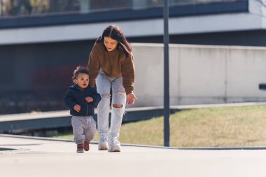 Genç esmer anne mahalledeki melez bebeğini kovalıyor ya da cesaretlendiriyor. İki kişilik aile..