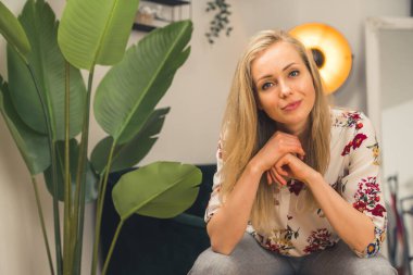 İyi giyimli, kendine güvenen beyaz, sarışın kadın yüzüne yakın avuç içleriyle koltukta otururken kameraya bakıyor. Modern dekor.