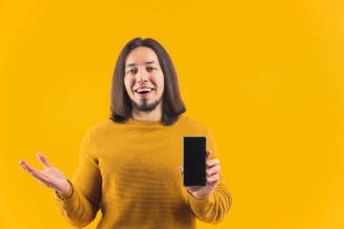 Heyecanlı genç adam kameraya siyah ekranlı akıllı telefonu gösteriyor ve başka bir el açık reklamı ve tanıtım konseptini tutuyor. Fotokopi alanı sarı arka plan çekimleri izole edildi. 