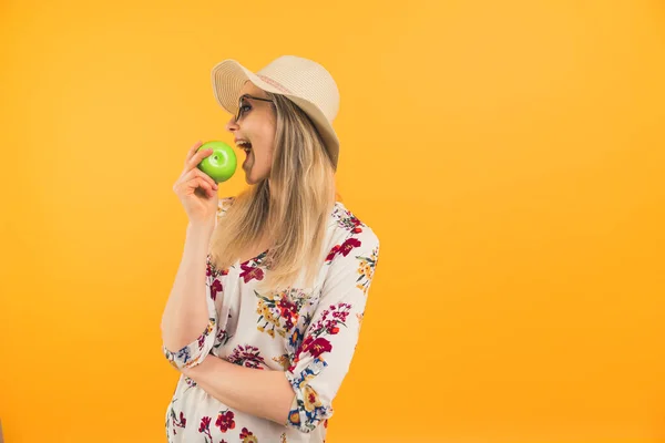 Young caucasian blond-haired woman in a straw hat eating fresh green apple. Medium studio shot over orange background. —  Fotos de Stock