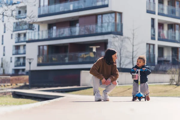 Weekend in the city. Young dedicated mother kneeling down and supporting her biracial little toddler boy who uses a scooter for the first time. — Stock fotografie