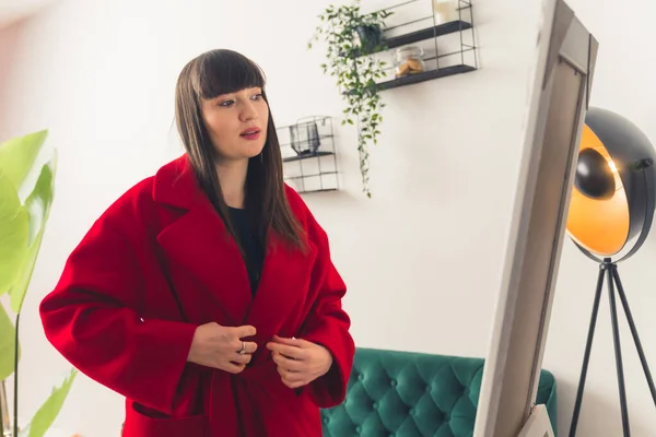 Conceito de compras online. Mulher de cabelos castanhos caucasiana na moda experimentando casaco vermelho quente na frente do espelho. — Fotografia de Stock