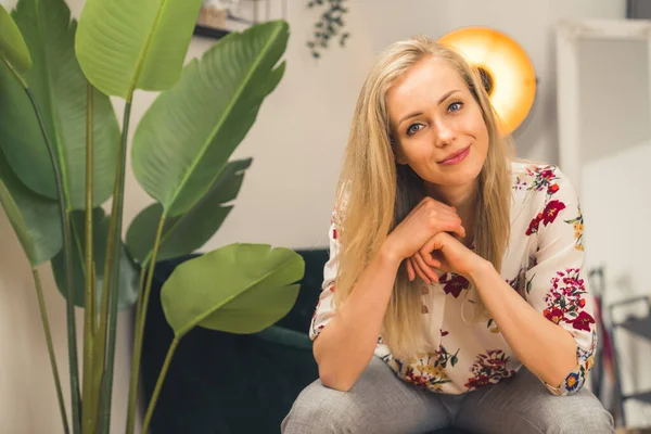 Beautiful young caucasian blonde woman sitting on a couch and looking at camera with positive facial expression. Modern flat interior. — Zdjęcie stockowe