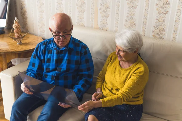 Pensioner caucasian spouses looking at x-ray results while sitting on a beige leather sofa. Support during illness. — Stockfoto