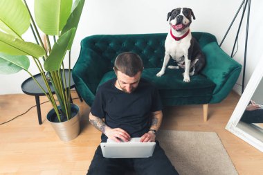 Beyaz dövmeli, siyah tişörtlü orta yaşlı bir adam yerde oturuyor ve büyük siyah beyaz köpeği kanepede otururken laptopunu kullanıyor. İç mimari.