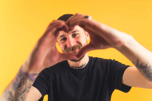 Tattooed European guy with a mustache and a beard wearing black t-shirt showing heart sign gesture with two palms. Studio shot over yellow background, — ストック写真