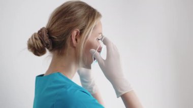Sarışın beyaz kadın doktor, beyaz arka plan üzerinde cerrahi maskeyi nasıl güvenli bir şekilde yerleştirip çıkaracağını gösteriyor..