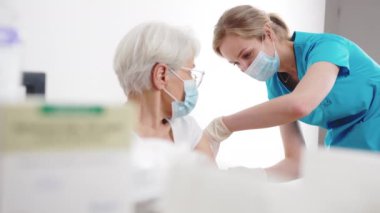 Virüs önleme. Genç, beyaz maskeli hemşire kadın yaşlı hastasını doktor muayenehanesinde aşılıyor..