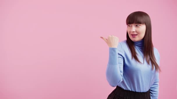 Young caucasian brunette woman wearing violet turtleneck showing copy space with her thumb over pink background. Medium studio shot. — ストック動画