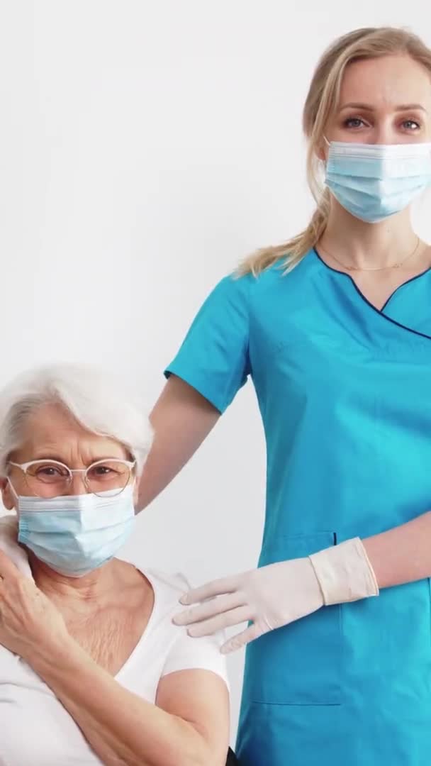 Vertical shot of two caucasian women - blonde nurse and elderly short-haired retired patient touching each others hands and looking at each other over white background. — Video