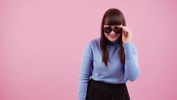 Funky caucasian brunette with bangs slowly takes off her sunglasses and laughs to the camera over pink background. — ストック動画