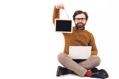 Bacak bacak üstüne atmış sakallı bir adam dizlerinin üstünde dizüstü bilgisayarını tutarak ipin üzerinde küçük siyah bir masa gösteriyor.