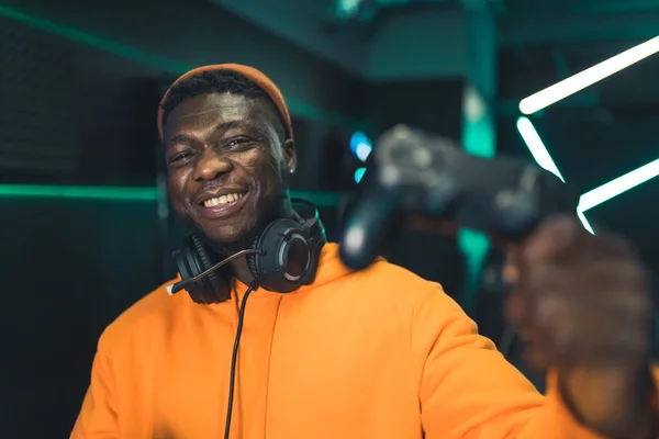 Satisfied smiley African-American gamer guy in orange hoodie looking at camera and smiling, while holding black game pad controller. — Fotografia de Stock