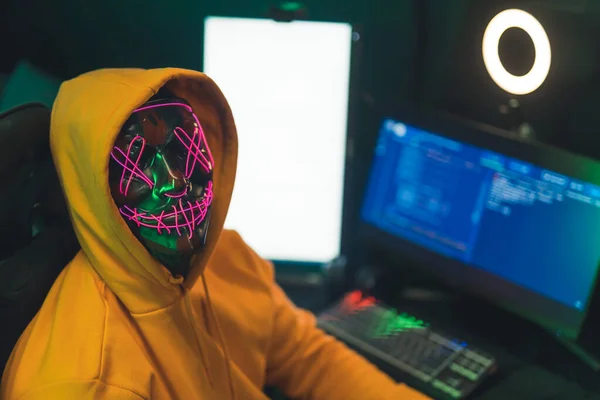 Concepto anónimo de identidad en internet. Persona en una sudadera con capucha amarilla delante de una computadora de programación que usa una espeluznante máscara de neón iluminada y mira a la cámara. —  Fotos de Stock