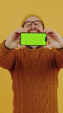 Portakal rengi arka planda izole edilmiş, boş ekranlı telefon gösteren neşeli genç adam dikey video stüdyosu çekimi.
