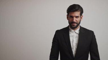 Siyah smokinli ve beyaz gömlekli baştan çıkarıcı sakallı Kübalı işadamı kollarını göğsünün önünde kavuşturuyor ve gri arka planda doğrudan kameraya bakıyor. Orta stüdyo çekimi..