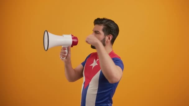 Knappe baard brunet man in de 30 in een t-shirt met Cubaanse vlag motief energiek schreeuwen via luidspreker over oranje achtergrond. — Stockvideo