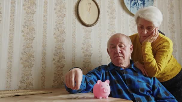 Mature Caucasian man puts coin into the piggy bank, wife standing beside savings concept — стоковое видео