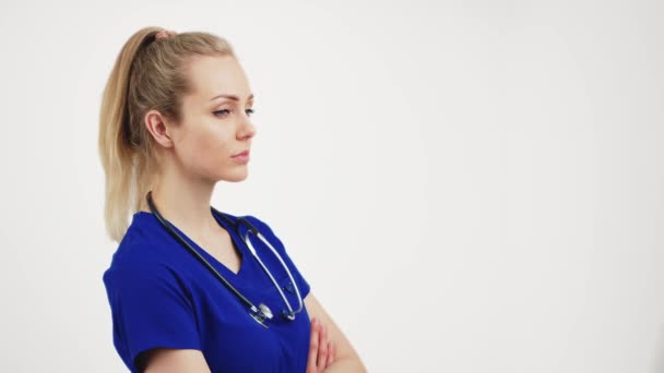 Blonde beautiful nurse stands facing the camera with her hands folded, isolated white background copyspace — Stockvideo