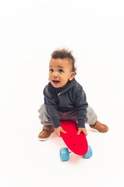 Kırmızı kaykayın üzerinde oturan şirin çocuk şaşırmış görünüyor, beyaz arka plan, stüdyo çekimi.