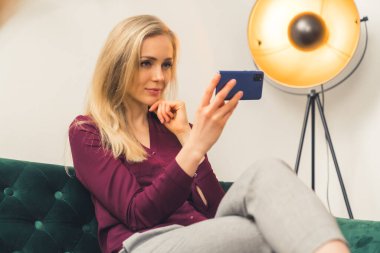 Beautiful young blond woman watching video on her smartphone sitting in the sofa copy space 
