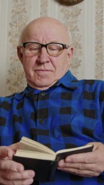 Senior white-haired Caucasian man in glasses reads book vertical video 