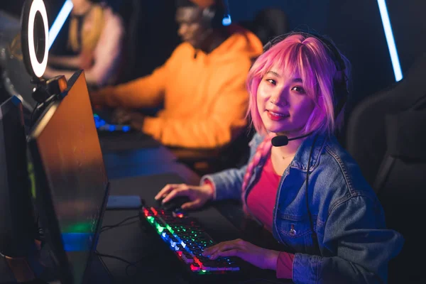 PC bang. Linda chica de pelo rosa asiática relajada mirando a la cámara mientras usa auriculares profesionales, y jugando en la computadora de escritorio. —  Fotos de Stock