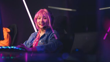 Boş zaman aktiviteleri konsepti. Pembe peruklu genç bayan oyuncu, kablosuz kulaklık kullanıyor ve renkli klavyesiyle kameraya bakıyor..