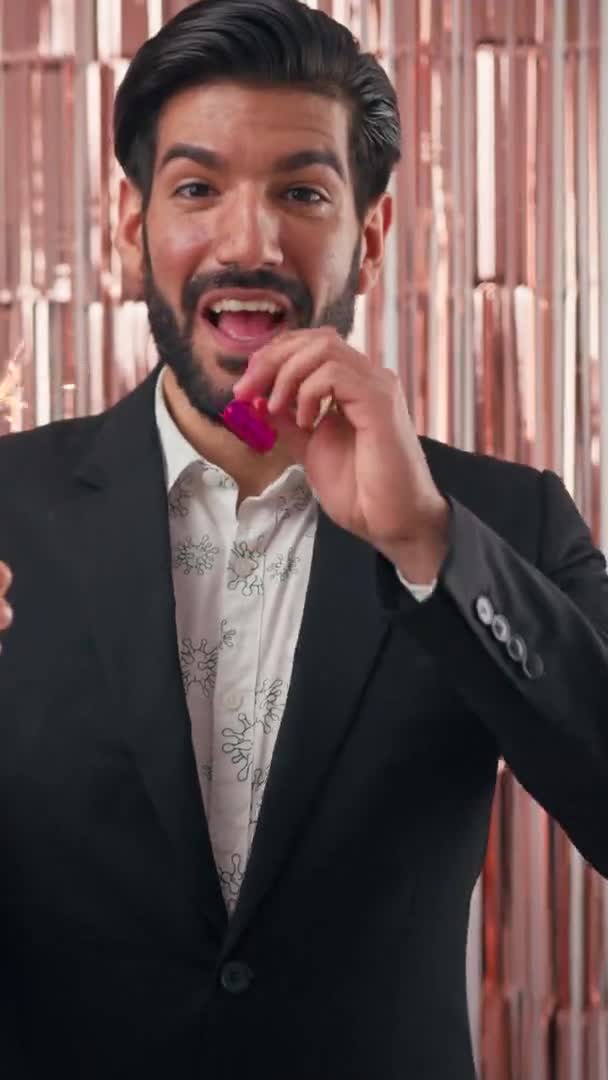 Chic handsome bearded Cuban man in his 30s in elegant suit celebrating New Years Eve or birthday, holding sparklers, and blowing party horn. Vertical studio shot. — Vídeo de Stock