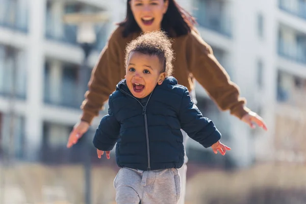 Mother running behind extremely happy joyful biracial toddler son on a path. Outdoor shot.